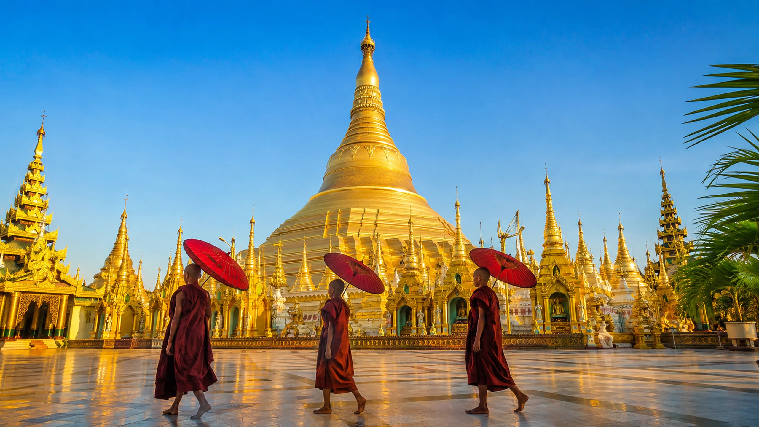 Three novice monks carrying red umbrellas across the Shwedagon Pagoda terrace in Yangon